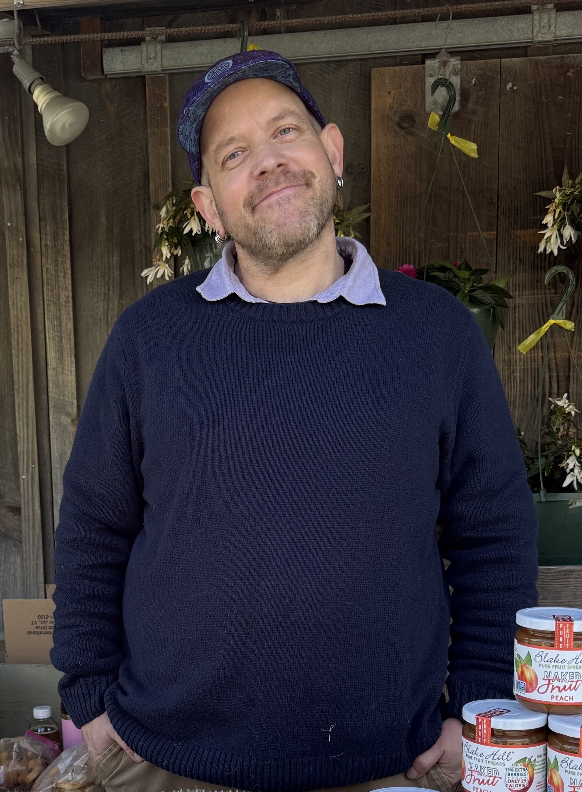 Jesse Barrows in a navy sweater smiles at a market stand with jars of fruit spreads.