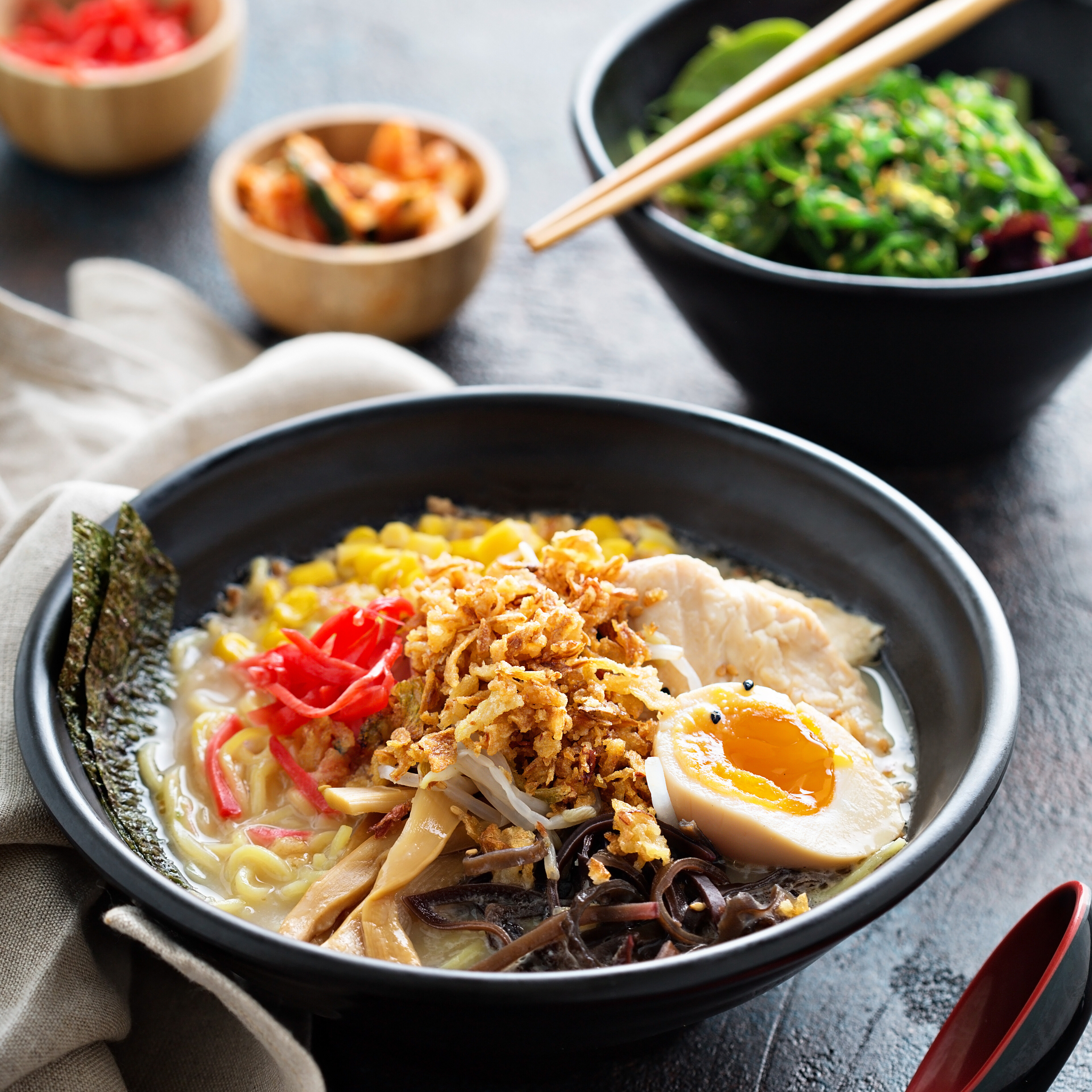 A bowl of ramen topped with chicken, egg, vegetables, and crispy onions, with sides.