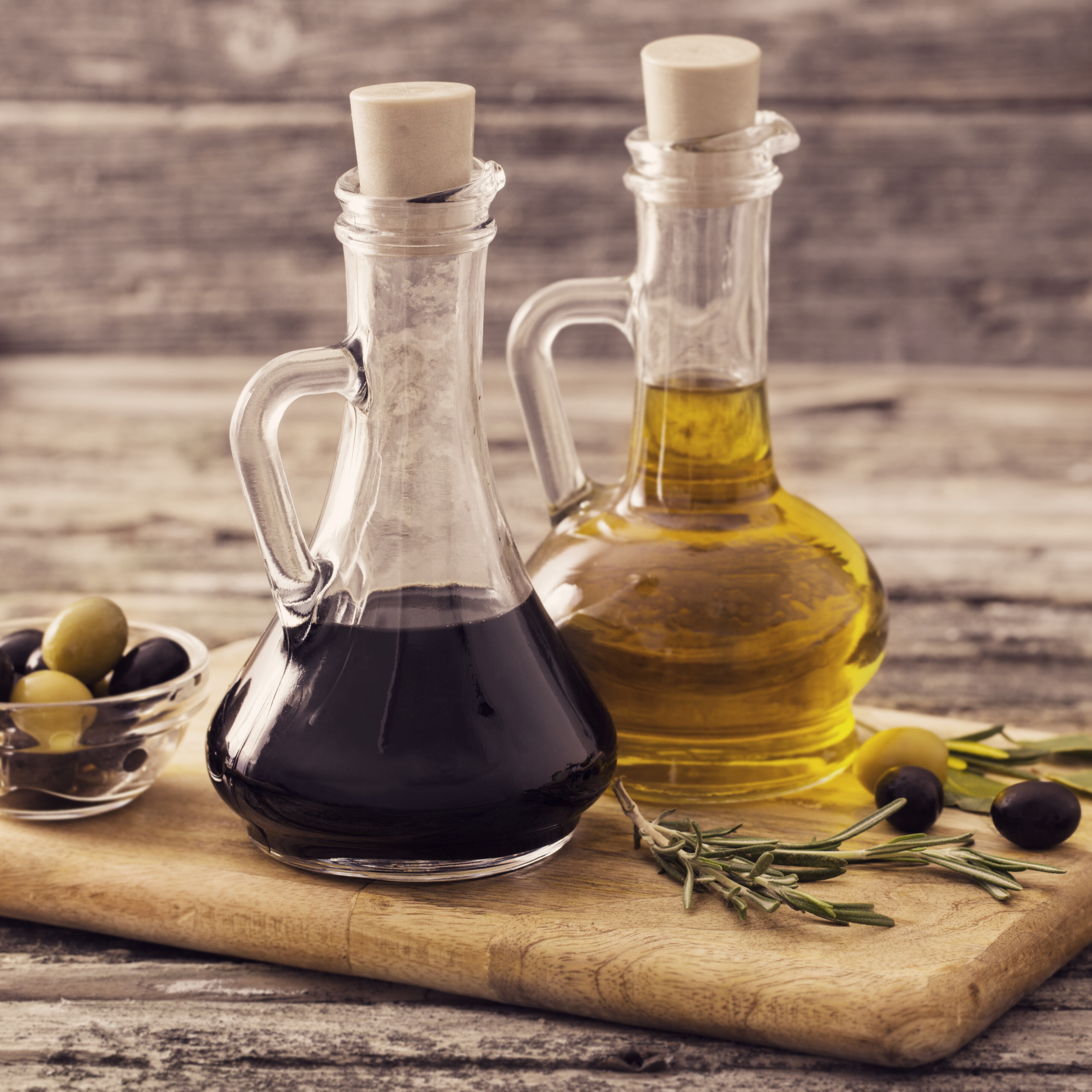 Glass bottles of olive oil and balsamic vinegar on a wooden board with olives.