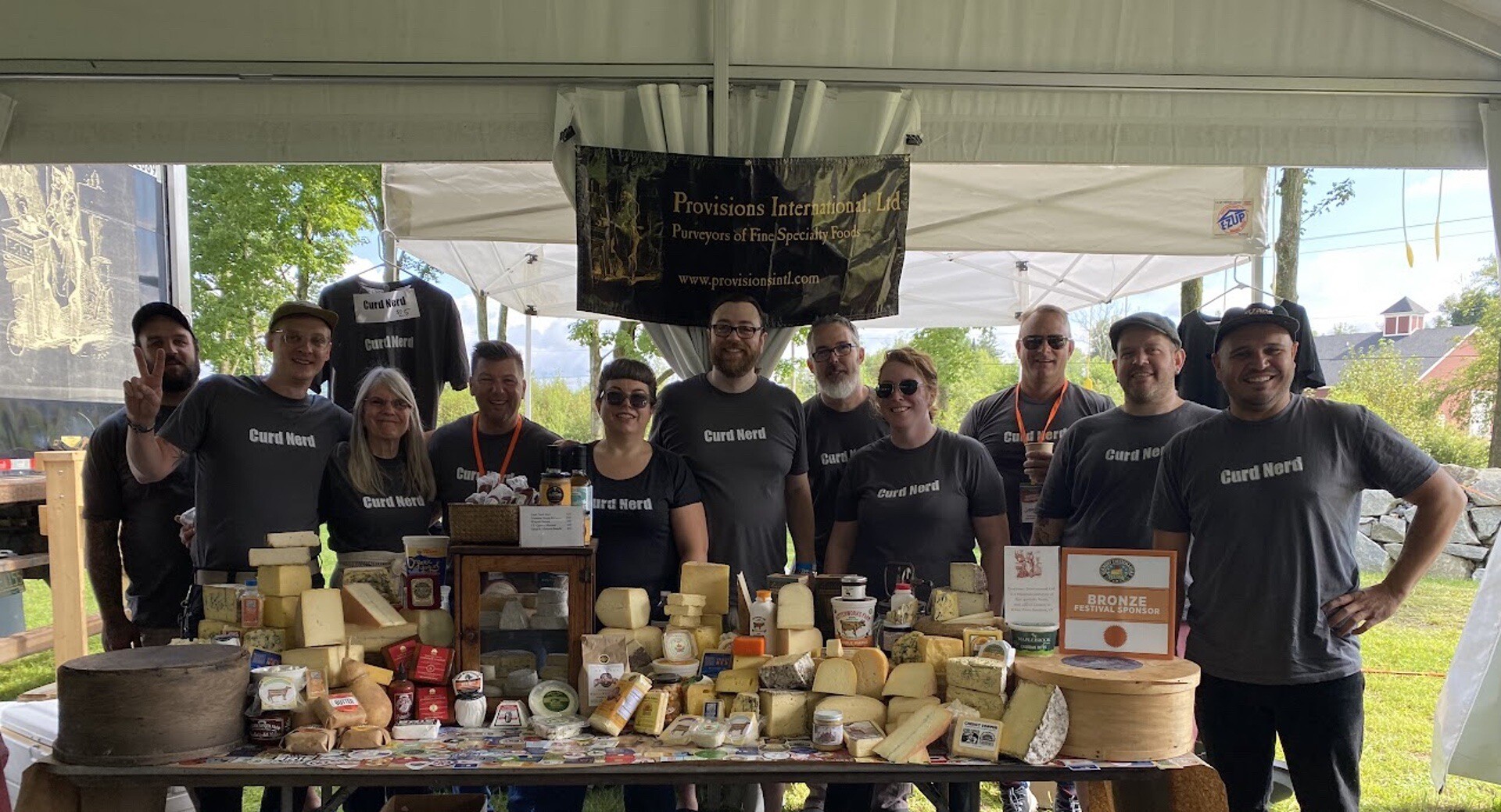 Provisions International staff in "Curd Nerd" shirts posing at a cheese display under a tent.