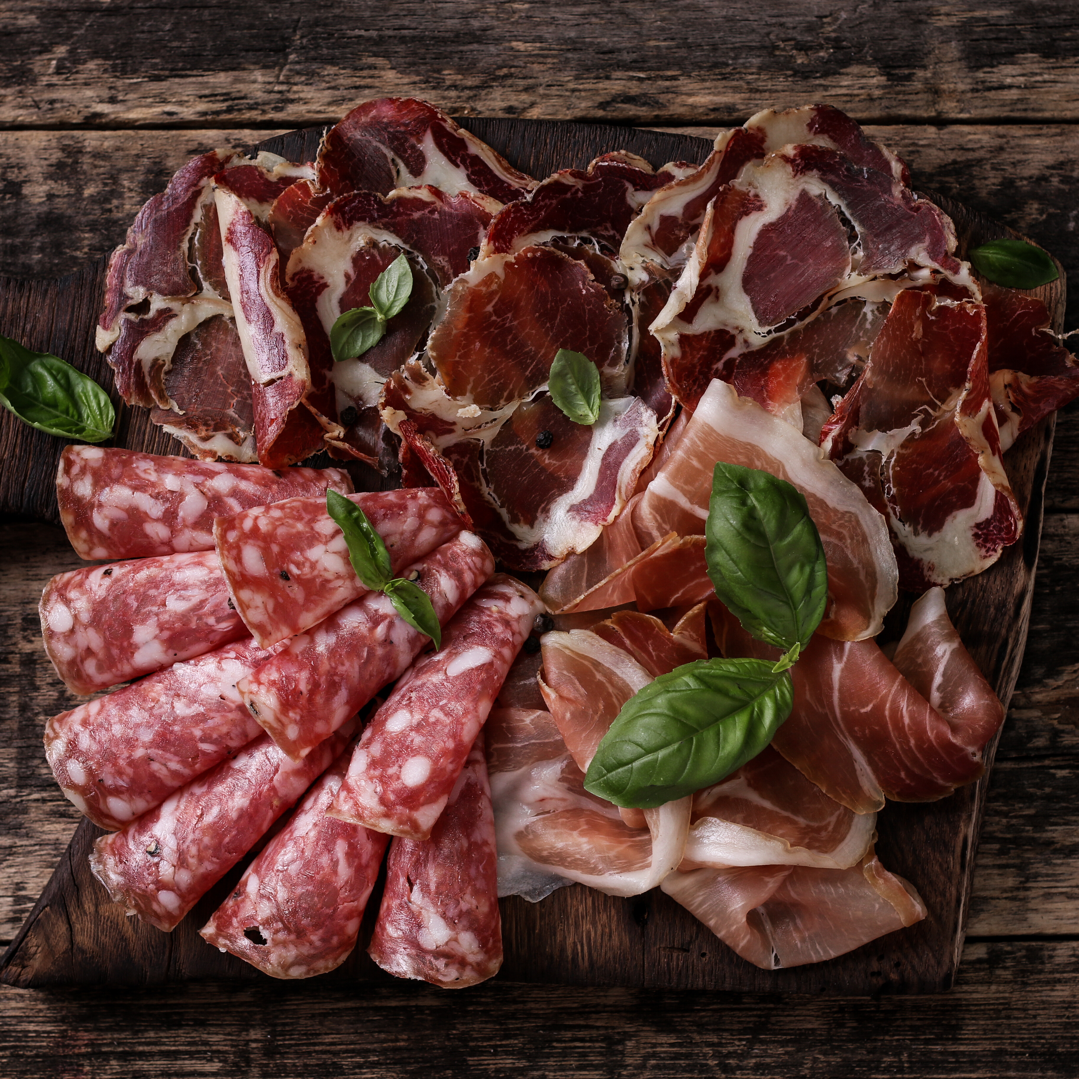 Assorted cured meats arranged on a wooden platter, garnished with basil leaves.