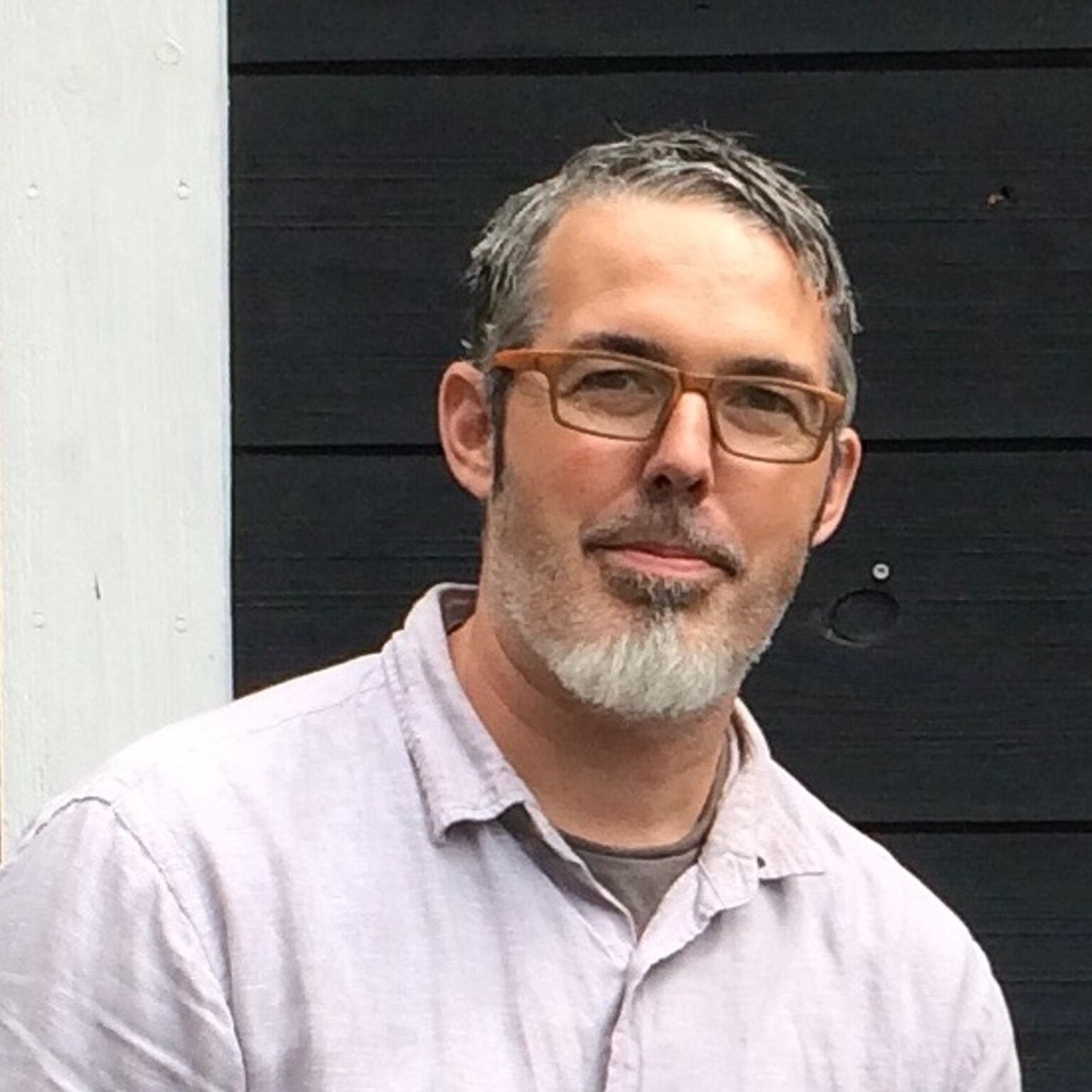 A man with glasses and a beard smiles in front of a black wooden background.