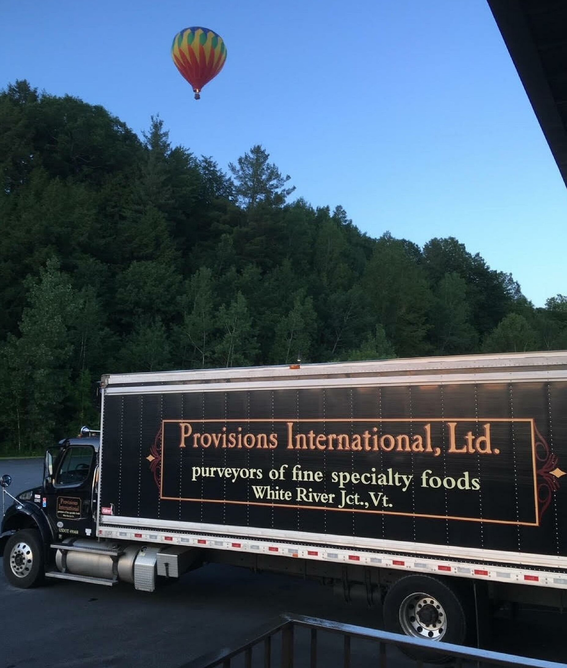 A colorful hot air balloon floats above a food delivery truck parked near trees.